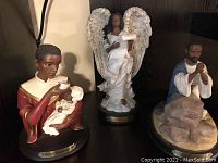 Three religious Ebony Treasures figurines displayed together; woman feeding baby, angel with wings, man praying on rock.