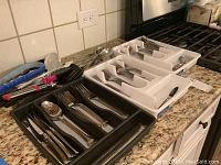 Wide view of several flatware trays containing knives, forks, spoons, and serving utensils along with kitchen utensils on granite countertop.