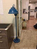 Full view of the vintage blue metal floor lamp standing beside a cabinet and near double doors, showing the overall height and structure including shade and base with decorative perforations.