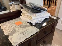 Lot stack including black packaged curtain swags, white and cream tablecloths on black marble surface