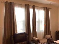 Four gold curtain panels and two white sheer panels hanging on a rod over two windows in a room with light walls and furniture including leather chair and armchair.