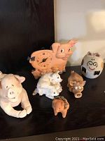 Photo showing six pig figurines on black surface, including vintage orange pig clock, two small brown pigs, white round pig, cream-colored pig with piglets, and a large light pink pig.