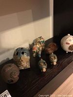Seven pig figurines on dark surface, mixed materials including ceramic and paper-mâché, varying sizes and colors.