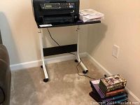 Front view of the adjustable metal printer stand with a printer placed on top (printer not included). Shows white metal frame and black pressboard top surface.