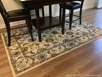 Full view of the cream decorative rug under a wooden table showing overall size, shape, and placement on wood floor.