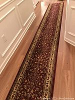 Long view of the burgundy runner rug laid out in a hallway showing its full length and beige borders with intricate floral designs.
