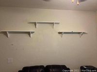 Three white wooden wall-mounted shelves arranged on a cream colored wall above a dark couch, showing overall condition and setup.