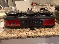 Front view of Bella triple slow cooker with three removable black crocks and glass lids on a granite countertop.