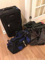 Photo showing one black soft-side rolling suitcase standing upright with four travel bags, including a blue and black duffel bag and three black bags of various sizes stacked or leaning beside it on a wood floor.