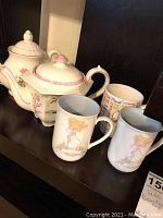Two white ceramic teapots with floral and pink details alongside five mugs on shelf; three mugs feature Precious Moments cartoon girl 'Dorothy' and two have text designs.