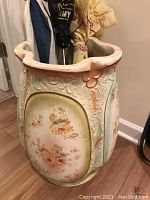 Front angled view of large ceramic umbrella stand showing ornate scrollwork and painted floral motif, with umbrella handles inside.