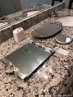 Photo of two vanity trays, hand mirror, and ceramic toothbrush holder arranged on granite countertop.