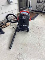 View of the full wet dry vacuum with hose attached and extension wand in use, showing the black cylindrical body with red handle and latches, on concrete floor.
