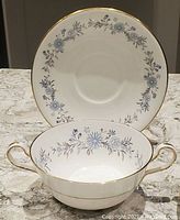 Photo showing a two-handle bone china bowl in front of a matching saucer, both decorated with the Aynsley Mayfield blue floral pattern and gold edging.