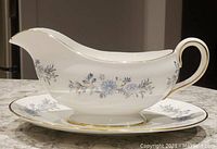 Side view of Aynsley bone china gravy boat with blue floral border and gold trim on white base, placed on matching underplate.