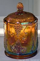 Full view of amber/marigold iridescent carnival glass lidded jar with the harvest grapes and leaves motif visible on lid and body.