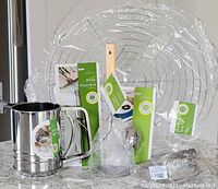 Photo showing six kitchen accessories including sifter with handle, peeler in original packaging, tea ball infuser, cooling rack, and dough blender all arranged on countertop.