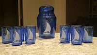 Full set photo showing 1 pitcher and 10 glasses on marble countertop. The blue glassware displays nautical sailboat imagery in white.