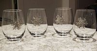 Four clear stemless wine glasses, each with an applied silver-tone snowflake decoration featuring faux sparkling jewels, arranged in a row on a marble surface.