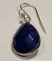 Close-up photo of front side of one earring showing deep blue faceted lapis lazuli teardrop in sterling silver bezel, with hook detail.