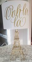 Full view of the lamp showing gold wireframe Eiffel Tower base and white fabric shade with 'Ooh la la' text.