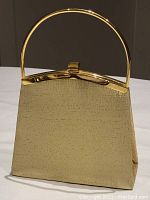 Front angled view of gold tone vintage Daniel Ames evening purse, showing texture of metallic fabric and handle attached to clasp frame.