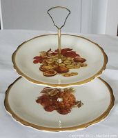 Front view of vintage 2-tier cake tray showing detailed fruit pattern on top and bottom plates, gold trim, and metal handle.
