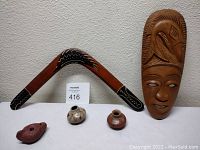 Photo of tall carved wood mask with bird headdress, Murra Wolka handpainted boomerang, red clay ocarina flute, and two small painted pottery vases arranged on white surface.
