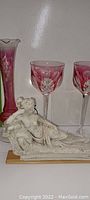 Small white partial nude figurine lying on rectangular base next to pink ruffle rim bud vase and two pink and clear wine glasses