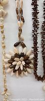 Close-up image showing a necklace with a large flower-shaped pendant made of alternating white and dark brown shells, alongside other single-strand dark brown shell necklaces.