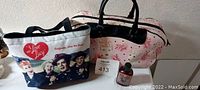 Photo shows two I Love Lucy handbags: one large pink bag with various Lucy faces and black handles, and one smaller tote with the classic cast photo of the show on white background, along with a small bottle of Vita-Meat-A-Vegamin on the side.