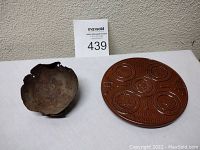 Two items: an old worn tin bowl with a rustic surface and a round brown pottery trivet embossed with tribal seals.