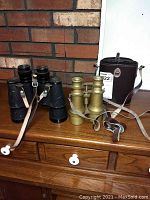 Two pairs of binoculars side by side on wooden dresser against brick wall background; one brass-colored pair with strap and one black textured pair with strap; dark brown carrying case behind brass pair.