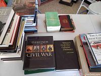View of multiple hardcover books stacked in piles on a table including the book 'Eyewitness to the Civil War', the dictionary, and other assorted books.