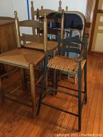 Two wooden rush seat bar stools, two black metal folding chairs, and one black wooden doll chair with woven seat shown together.