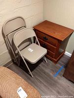 Two beige metal folding chairs next to a wooden side table with three drawers, placed against a beige wall on a concrete floor.
