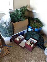 Boxed and loose artificial Christmas tree branches, pine garland in bags and box, wrapped wreath, and ceramic Santa statue boxes on floor.