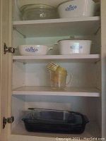 Cupboard with two shelves holding Corning Ware dishes with blue cornflower design on top and middle shelves, plastic measuring cups with plastic forks on middle shelf, and large clear Pyrex glass casserole dish on bottom shelf.