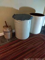 Three ceramic crocks side by side; two large crocks and one smaller butter churner with wooden plunger stick