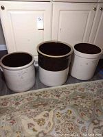 Three ceramic crocks placed side by side on floor against cabinet, showing varying sizes and glaze colors.