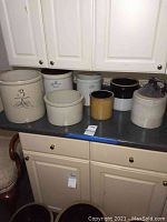 Photo of seven ceramic crocks of various sizes on countertop with white cabinets in background