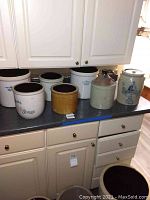 Seven ceramic crocks arranged on a kitchen counter, showing variety in size and markings.