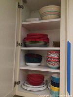 Kitchen cabinet shelves displaying stacked bowls and plates organized by color and material, including multicolor striped mixing bowl on top shelf, red and teal plastic bowls, and colorful patterned Euro ceramic bowls with chips.