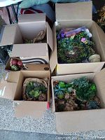 Four open boxes containing assorted artificial succulents, flowers, wreaths, and faux fruit showing overall lot contents