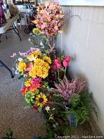 Photo showing multiple faux floral arrangements including sunflowers, tulips, dahlias, branches, and ferns in pots and baskets lined up near a wall.