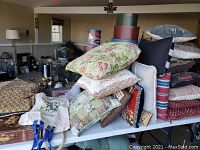 Photo of table with a pile of decorative pillows, chair cushions, some folded textiles and nesting boxes in background.