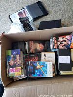 Box containing assorted VHS tapes, DVDs and CDs in cases, visible titles and genres.