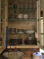 Cabinet interior showing stacks of white china plates, cups, and bowls with blue and gold trim; top shelf with glass stemware.