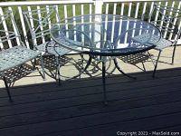 Wide view of glass top round patio table with three matching metal lattice chairs on outdoor deck.