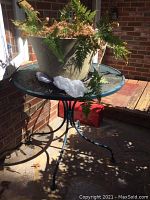 Full view of metal patio table with beige planter and fern plant placed on top, showing the table legs and shadow on concrete surface.